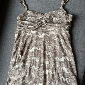 Brunmönstrad linne med smala axelband - Supersött linne med smala axelband och brunt och vitt blommigt mönster. Linnet har rynkade detaljer vid bysten och är tillverkad i ett mjukt, stretchigt material som känns skönt mot huden. Perfekt för varma dagar när du vill känna dig både bekväm och snygg.