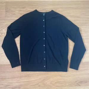 Svart kofta från Uniqlo, storlek L - En klassisk svart kofta från Uniqlo i storlek L. Koftan har rund halsringning, långa ärmar och knäppning med svarta knappar framtill. Tillverkad i mjukt material som känns skönt mot huden. Perfekt att slänga över en t-shirt eller skjorta.