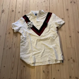 Chief keef polo  - Vit pikétröja från Polo Ralph Lauren med svart och vinröd V-formad detalj över bröstet, broderad logga och emblem på vänster sida. Klassisk krage, korta ärmar och slim fit. Perfekt för dig som gillar sportig och preppy stil.