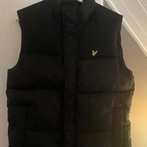 Svart dunväst från Lyle & Scott - Svart dunväst från Lyle & Scott med klassisk gul broderad logga på bröstet. Västen har hög krage och quiltad design som ger en schysst siluett. Perfekt att slänga över en hoodie eller tröja när det är kyligt ute.