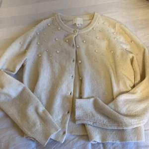Beige kofta med pärlor från H&M - Superfin beige kofta från H&M med pärldetaljer över axlar och bröst. Koftan har rund hals, ribbade muddar och knäppning framtill. Perfekt för dig som vill ha en söt och trendig look med extra detaljer.