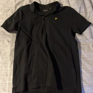 Pikétröja från Lyle & Scott - Svart pikétröja från Lyle & Scott med krage och korta ärmar. Tröjan har en gul broderad logga på bröstet. Tillverkad i mjuk bomull som är skön att bära hela dagen. Perfekt stil för sommaren och i top skick🔥