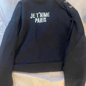 Svart sweatshirt med text Je t'aime Paris - Svart sweatshirt med broderad vit text 'JE T'AIME PARIS' på bröstet. Tröjan har rund halsringning, långa ärmar och dekorativa knappar på ena axeln. Perfekt för dig som gillar enkel men snygg stil.