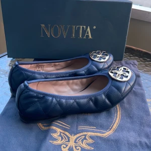 Mörkblå ballerinaskor från Novita - Snygga mörkblå ballerinaskor från Novita med rund tå och platt sula. Skorna har en stor guldfärgad metallogo framtill som ger en lyxig touch. Perfekta för dig som gillar klassisk men trendig stil. Köpa för nypris 1100kr, testat hemma men är oanvända. 