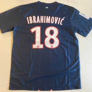 PSG Ibrahimovic 18 matchtröja Nike - Paris Saint-Germain fotbollströja replika med Ibrahimovic och nummer 18 på ryggen. Kolla bilder för defekter. Djupblå med vita och röda detaljer, klassisk PSG-design. Nike Dri-FIT-material som andas, kortärmad med klubbmärke och sponsortryck på bröstet. Perfekt för fans av Zlatan och PSG. Handtvätt endast!