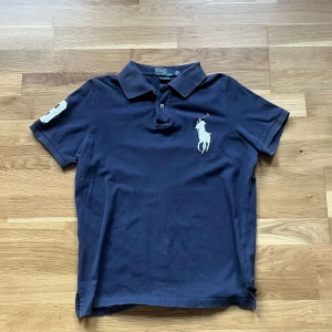 Ralph lauren piké  - Snygg marinblå t-shirt från Polo Ralph Lauren med klassisk krage och korta ärmar. Stor vit broderad logga på bröstet och detalj på ärmen. Tillverkad i mjuk bomull, perfekt för en clean och sportig look.