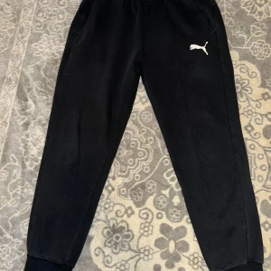 Svarta mjukisbyxor från Puma - Svarta mjukisbyxor från Puma med klassisk logga på låret. Byxorna har resår i midjan och vid bensluten, samt sidofickor. Tillverkade i mjukt material som är perfekt för chill eller träning. Enkel och stilren design som funkar till det mesta.