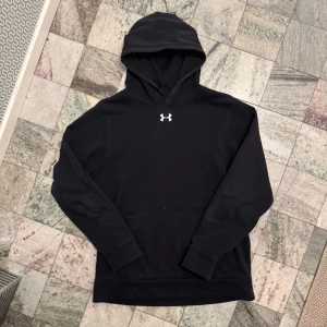Svart hoodie från Under Armour - Säljer en svart hoodie från Under Armour med deras logga på bröstet. Den har en bekväm passform och är perfekt för kyligare dagar tack vare ColdGear-materialet. Hoodien har en stor ficka framtill och långa ärmar.