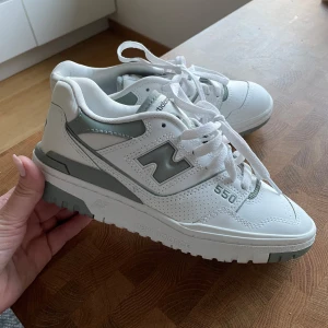 New Balance 550 vita sneakers - Snygga New Balance 550 sneakers i vitt med grå detaljer. Klassisk chunky siluett med perforerade partier och tydlig logga på sidan. Skorna har snörning och platt sula, tillverkade i skinn och syntet. Helt nya och säljer för att jag inte hann returnera i tid pga fel storlek. Storlek 36.5
