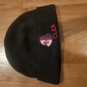 Svart Moncler mössa med broderad logga - Svart mössa från Moncler med uppvikt kant och broderad logga i rött, blått och vitt framtill. Tillverkad i 100% akryl, perfekt för kalla dagar. Snygg och stilren accessoar med tydlig Moncler-text på kanten.