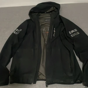 Svart Gore-Tex vindjacka Sail Racing - Svart vindjacka från Sail Racing med huva och snygga logodetaljer på ärmar och rygg. Jackan är tillverkad i slitstark Gore-Tex och har dragkedja framtill. Insidan har coola oranga detaljer och Sail Racing Link Technology för extra funktion.