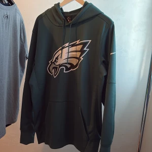 Grön Philadelphia Eagles hoodie Nike - Snygg mörkgrön hoodie från Nike med Philadelphia Eagles-logga på bröstet. Tröjan har huva med snörning, magficka och långa ärmar. Perfekt för dig som gillar amerikansk fotboll och vill ha en sportig stil.