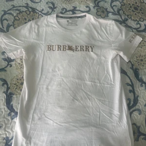 Vit Burberry t-shirt  - Vit t-shirt från Burberry i bomull med klassisk rund hals och korta ärmar. Stor Burberry-logga i brunt och guld framtill samt mindre logga på ena ärmen. Enkel och stilren design som passar perfekt till jeans eller shorts.