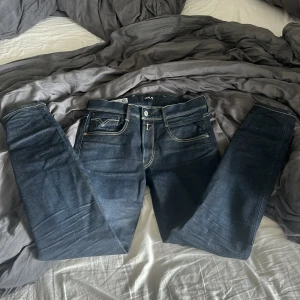 Replay Anbass mörkblå jeans 31/32 - Snygga Replay Anbass jeans i mörkblå tvätt med kontrastsömmar och klassisk femficksdesign. Byxorna har raka ben och normal passform, med diskret Replay-logga på fickan och coola detaljer på bakfickorna. Perfekta för dig som gillar stilrena jeans med lite edge.