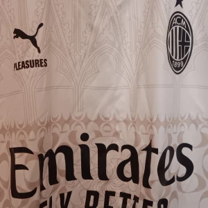 AC Milan x Puma vit fotbollströja - AC Milan fotbollströja i vitt med snygga grafiska detaljer inspirerade av gotisk arkitektur. Puma-logga, klubbmärke och flera sponsorer tryckta i svart. Svartmönstrade muddar och lätt, ventilerande material. Perfekt för dig som älskar italiensk fotboll. Perfekt för sommar.