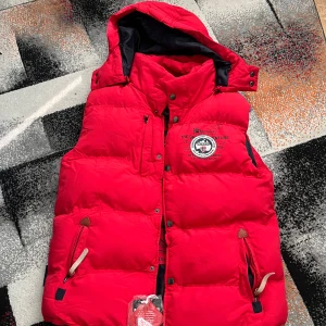 Röd dunväst från Geographical Norway - Riktigt fet röd dunväst från Geographical Norway med svart innerfoder och stora tryck både inuti och utanpå. Västen har huva, dragkedja och knappar framtill samt flera fickor med snygga detaljer. Perfekt för kalla dagar när du vill sticka ut.