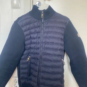 Mörkblå Moncler Cardigan  - Snygg mörkblå pufferjacka från Moncler med quiltad front och stickade ärmar. Jackan har dragkedja framtill, två fickor med dragkedja och klassisk Moncler-logga på ärmen. Perfekt för dig som vill ha en stilren och sportig look.