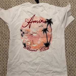 Oversize Amiri tshirt - Vit t-shirt från Amiri med tryck i rosa och svart på ryggen som visar en strand med palmer och surfare. Framsidan har en mindre Amiri-logga i rött och rosa. Klassisk passform och korta ärmar, perfekt för en chill sommarlook.