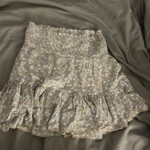 Beige blommig volangkjol från 157 - Supersöt kort volangkjol från 157 i beige med vitt blommönster. Kjolen har smock i midjan för en bekväm passform och flera lager volanger som ger en flowy känsla. Perfekt för vår och sommar när du vill ha en chill och feminin look. 