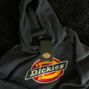 Mörkblå Dickies hoodie med logga - Snygg mörkblå hoodie från Dickies i storlek medium. Stor Dickies-logga i rött, gult och vitt framtill. Klassisk huva med snörning och känguruficka. Mjuk bomullskänsla och ribbade muddar vid ärmslut och nederkant.