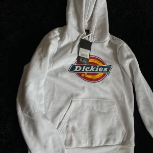 Vit hoodie från Dickies med logga - Snygg vit hoodie från Dickies i bomull med stor färgglad logga på bröstet. Klassisk känguruficka, huva med snörning och relaxed passform. Perfekt för dig som gillar streetwear och vill ha en enkel men stilren look.