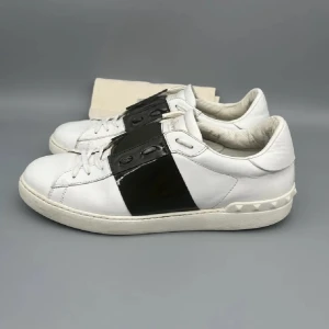 Valentino Garavani Open sneakers svart/vit - Snygga Valentino Garavani Open sneakers i vitt skinn med bred svart läderdetalj över snörningen. Klassisk rund tå, platt sula och ikoniska nitar baktill. Perfekta för dig som vill ha en clean men ändå edgy look.