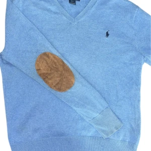 Blå stickad cardigan från Polo Ralph Lauren - Snygg ljusblå stickad tröja från Polo Ralph Lauren med v-ringning och klassisk logga på bröstet. Tröjan har långa ärmar och bruna mockadetaljer på armbågarna för en extra touch. Perfekt för dig som gillar stilren och tidlös design.