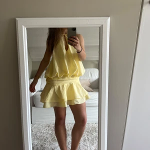 Gul volangklänning med halterneck - Supersöt ljusgul kort klänning med volangkjol och halterneck. Klänningen har en lös passform upptill, markerad midja och är ärmlös. Perfekt för varma dagar och ger en fräsch sommarkänsla. Klänningen är öppen i ryggen!