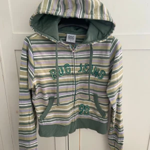 Randig hoodie från BDG Urban Outfitters - Säljer en randig hoodie från BDG Urban Outfitters i grönt, beige, vitt och lila. Tröjan har dragkedja, huva med snörning och broderad text 'BDG Jeans 98' framtill. Mjuk bomullskänsla och långa ärmar, perfekt för chill dagar. Har ett litet defekt under luvan men det märks inte. 