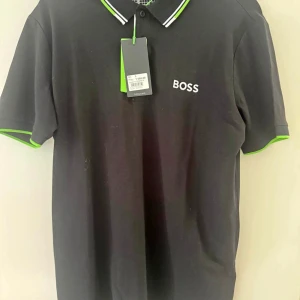 Svart pikétröja från BOSS med gröna detaljer - Snygg svart pikétröja från BOSS med vit logga på bröstet och fräscha gröna detaljer på krage och ärmslut. Klassisk pikékrage med knappar och vit rand. Perfekt för en stilren look och passar till många outfits.   HELT NY