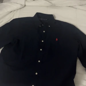 Mörkblå skjorta från Ralph Lauren M - Klassisk mörkblå skjorta från Ralph Lauren i bomull med knappar framtill och ikoniska röda broderade loggan på bröstet. Skjortan har lång ärm och button down-krage, perfekt för en clean och stilren look. Jag köpte den föra veckan för att jag skulle ha den på min brors bröllop och det blev inte det så jag vill säga den och jag har inte använt den 