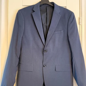 Blå kostym i polyester och viskos - Snygg blå kostym med klassisk kavaj och matchande byxor. Kavajen har två knappar, smala slag och fickor med lock. Materialet är en mix av polyester, viskos och lite elastan för extra komfort. Perfekt för dig som vill ha en stilren och modern look. Vit skjorta från H&M ingår (S). Använd 2 ggr, sen utväxt. 