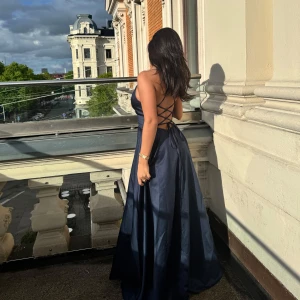 Marinblå långklänning med snörning - Elegant marinblå långklänning med öppen rygg och snygg snörning baktill. Klänningen har smala axelband, v-ringad omlott framtill och ett svepande, utsvängt fall. Perfekt för bal eller festliga tillfällen. Materialet har en lätt glansig finish som ger en lyxig känsla.