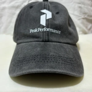 Grå Peak Performance keps - Snygg grå keps från Peak Performance med böjd skärm och justerbart spänne bak. Kepsen har vit logotyp och text framtill. Tillverkad i mjuk bomull som ger en avslappnad vibe. Perfekt accessoar för en sportig och stilren look.