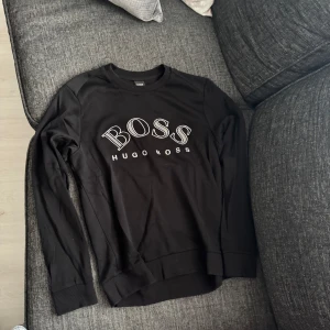 Svart långärmad tröja Hugo Boss - Svart långärmad tröja från Hugo Boss med stor BOSS-logga framtill i vitt. Tröjan har rund halsringning, ribbade muddar och en diskret dekorativ söm på ryggen. Perfekt för dig som gillar stilrena och klassiska plagg med en modern touch.