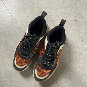 Chunky leopard sneakers från NA-KD - Chunky sneakers från NA-KD med leopardmönster i brunt och orange, svarta och vita detaljer samt grov platt sula. Skorna har snörning och cool streetstyle-vibe. Perfekta för dig som vill sticka ut med ett djurmönstrat statement.