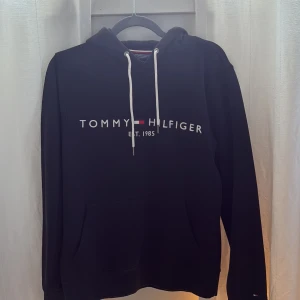 Svart hoodie Tommy Hilfiger S - Svart hoodie från Tommy Hilfiger med vit logga och brodyr framtill. Klassisk känguruficka, vita snören i huvan och mjukt material. Perfekt för chill dagar och snygg streetstyle. Säljes i storlek S.