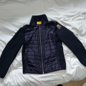 Mörkblå Parajumpers pufferjacka YS - Snygg mörkblå pufferjacka från Parajumpers med quiltad framsida och släta ärmar. Jackan har dragkedja framtill, hög krage och en ikonisk patch på axeln. Perfekt för dig som vill ha en stilren och sportig look.