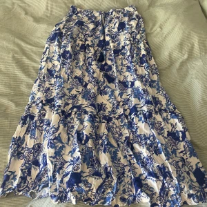 Blommig långkjol ifrån Lindex  - Superfin långkjol från Lindex med blått och vitt blommönster. Kjolen har volanger och avslappnad passform, perfekt för sommarens alla tillfällen. Tillverkad i ett lätt och svalt material som känns skönt mot huden.