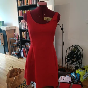 Röd ärmlös kortklänning - Snygg röd klänning med breda axelband och rundad urringning både fram och bak. Modellen är ärmlös och har en utställd kjol som ger en skön och ungdomlig siluett. Perfekt för dig som vill sticka ut med färg! Materialet känns mjukt och följsamt. Finns en liten fläck. Se bild!!