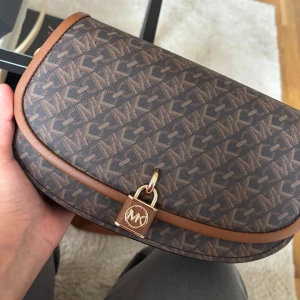 Brun axelväska från Michael Kors - Snygg brun axelväska från Michael Kors med MK-logga över hela väskan. Väskan har guldfärgade detaljer, justerbar axelrem i textil och skinn, samt ett lås framtill. Invändigt finns dragkedjefack och ljusbrunt foder med logotyp. (Pris kan diskuteras)