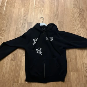 Svart hoodie med tryck från Bvaldi - Svart hoodie från Bvaldi med vita tryckdetaljer framtill och texten 'GUARDIANS' på ryggen. Tröjan har dragkedja, huva och två fickor framtill. Perfekt för en avslappnad stil.