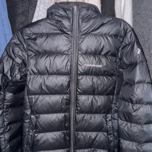 Svart dunjacka från Peak Performance - Säljer en svart dunjacka från Peak Performance med huva och dragkedja framtill. Jackan är quiltad och har en sportig look, perfekt för kyliga dagar. Klassisk design med diskret logga på bröstet.