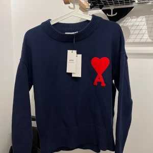 Mörkblå stickad tröja från Ami - Säljer en mörkblå stickad tröja från Ami med stor röd hjärtapplikation och bokstaven A på bröstet. Tröjan har rund halsringning och långa ärmar. Perfekt för dig som vill ha en stilren men ändå unik look.