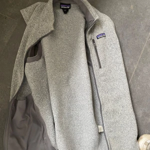 Grå väst från Patagonia - Säljer en ljusgrå fleeceväst från Patagonia med hög krage och dragkedja framtill. Västen har en bröstficka med dragkedja och är perfekt för lager-på-lager. Klassisk och stilren design.