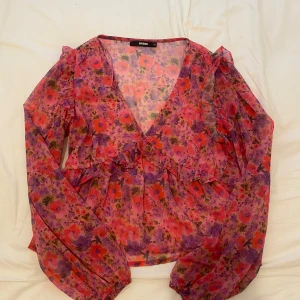 Blommig blus med volang från Bik Bok - Säljer en blommig blus från Bik Bok i rosa, lila och röda toner. Blusen har långa ärmar, v-ringning och volangdetaljer längs axlar och byst. Tunt och luftigt material, perfekt för vår och sommar.