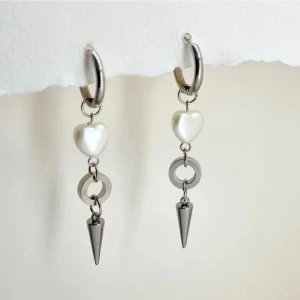 Silvriga örhängen med pärlhjärta och kon - Unika örhängen i silverfärg med hängande detaljer: ett vitt pärlhjärta, en rund länk och en spetsig kon. Perfekt för dig som vill sticka ut med en edgy och söt stil.Handgjorda örhängen. Material- 100% rostfritt stål ,plastik .Örhängeskrokar -rostfritt stål. Nickel fri. Längd -6,5cm. 