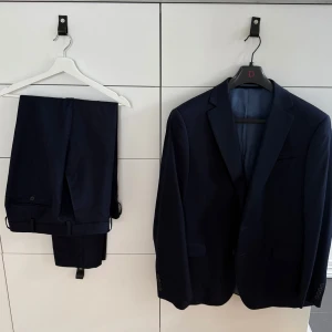 Mörkblå kostym från Dressmann - Säljer en klassisk mörkblå kostym från Dressmann i storlek 56 regular. Kavajen har två knappar och byxorna har storlek 40-32. Perfekt för formella tillfällen och stilrena outfits.