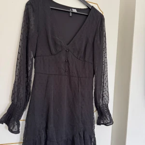 Svart långärmad klänning med prickigt mönster från H&M Divided - Säljer en svart långärmad klänning från H&M Divided med transparenta ärmar och diskret prickigt mönster. Klänningen har v-ringning, knappar framtill och volang vid ärmslut och nederkant. Perfekt för dig som gillar stilrena detaljer.