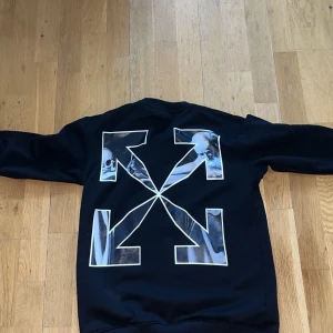 Svart sweatshirt från Off-White med tryck - Svart sweatshirt från Off-White med stort grafiskt tryck i vitt och blått på ryggen och mindre logga framtill. Rund halsringning och långa ärmar. Klassisk streetwear-stil med mjukt bomullsmaterial.